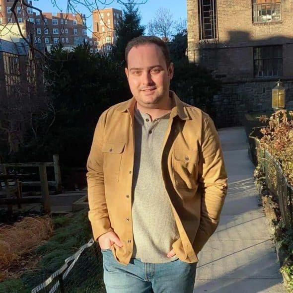 A man wearing a tan jacket over a gray shirt and blue jeans standing outside.