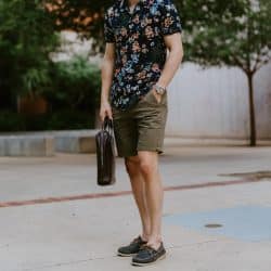A man standing outdoors wearing a floral short-sleeved shirt, olive shorts, and boat shoes, holding a bag.