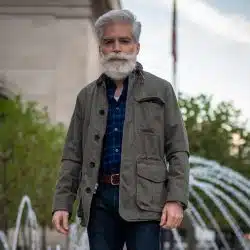 An man with a white beard and hair, wearing a gray-green field jacket over a blue plaid shirt and jeans, stands outdoors near a fountain.
