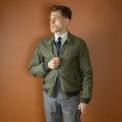 A man wearing a white shirt, a dark tie, gray trousers, and an olive green lightweight jacket with a zipper partially undone.