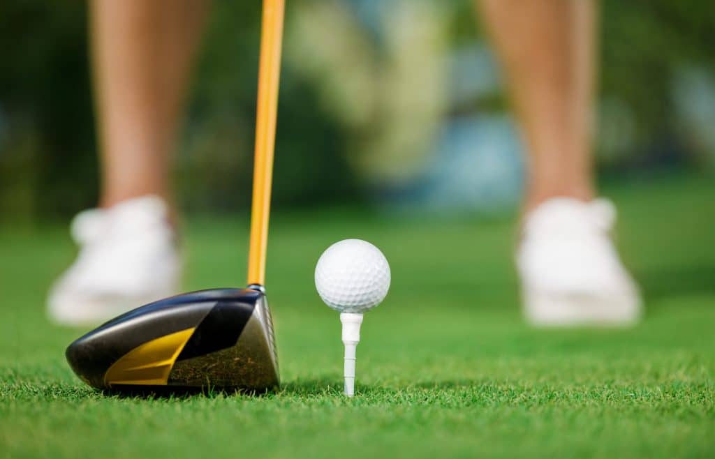 Close-up view from behind a golf ball on a white tee with a driver club head, showing the blurred legs and shoes of a golfer in the background.