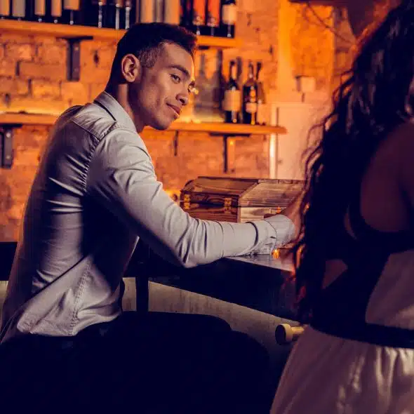 Smiling man at a bar looking towards a woman sitting across him.