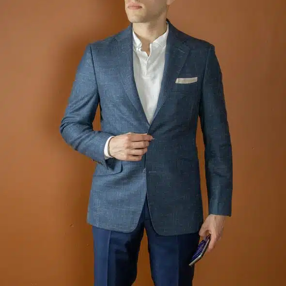 A man standing against a brown background, wearing a blue blazer over a white shirt and blue pants holding a phone in his left hand.