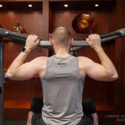 A man with a shaved head is lifting a barbell while wearing a gray tank top with the word "BARBELL" printed on it.
