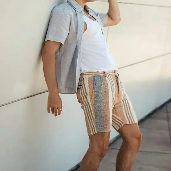 A man casually leaning against a white wall, wearing a light gray open shirt over a white tank top and striped shorts.