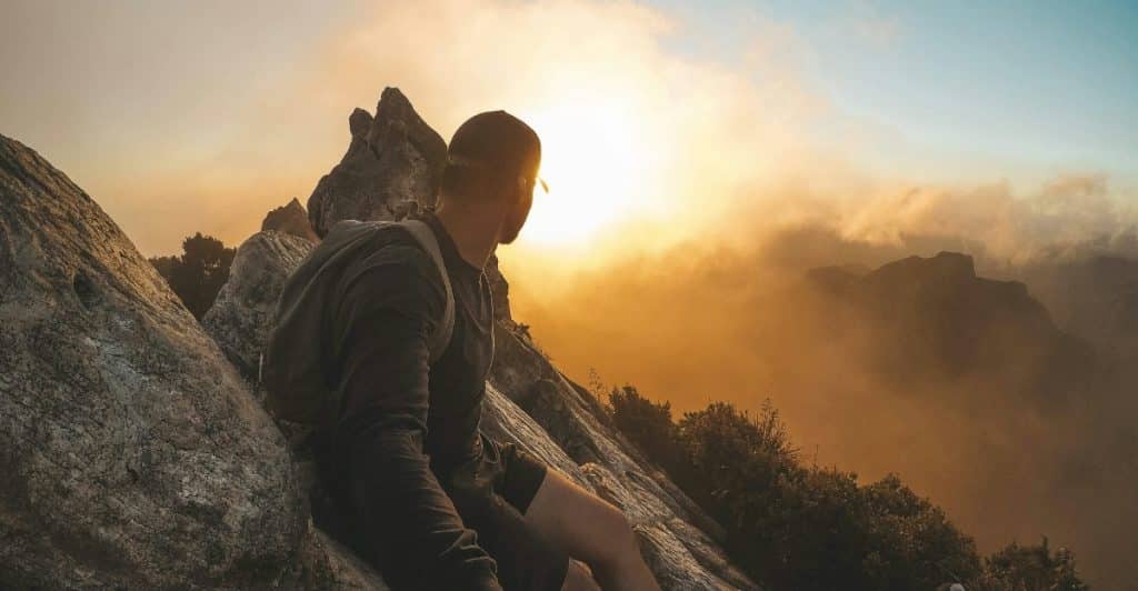 A person sitting on a rocky outcrop looking at a sunset over a mountainous landscape.