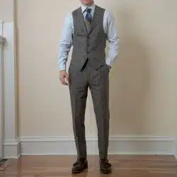 A man wearing a brown tweed suit vest and pants with a light blue shirt, patterned tie, and brown monk strap shoes.