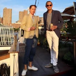 Two men standing on a boat deck at sunset, one in a tan blazer and blue pants, the other in a navy blazer and white pants.