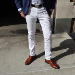 A man's lower body wearing white jeans and brown monk strap shoes, with a striped shirt and dark jacket visible.