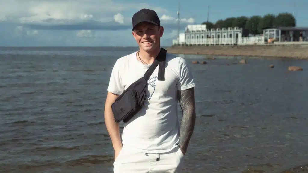 A man wearing a black cap, white clothes, and a shoulder bag stands on a beach in front of the ocean with his hands in his pockets and smiles.