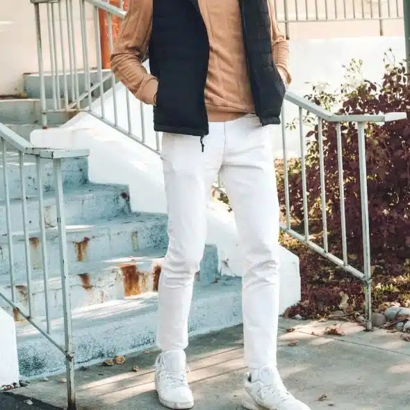 A man standing on outdoor stairs, wearing a black puffer vest, a tan long-sleeved shirt, white pants, and white sneakers.