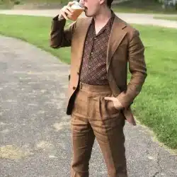 A man standing outside, holding a glass of beer, wearing a brown suit with a patterned shirt underneath.