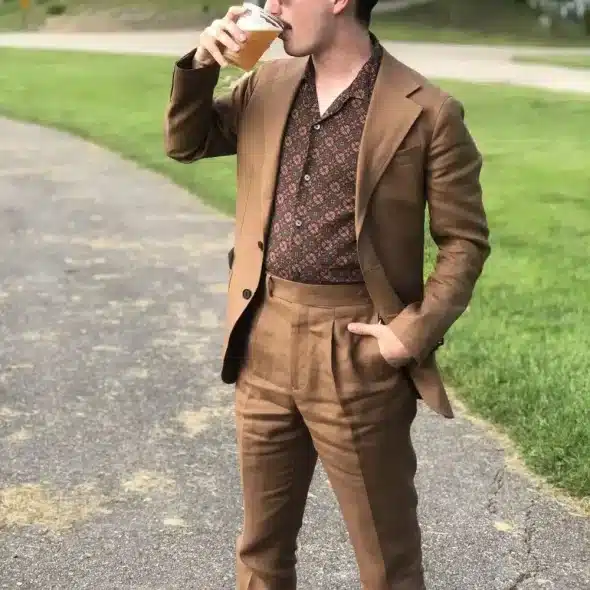 A man standing outside, holding a glass of beer, wearing a brown suit with a patterned shirt underneath.