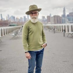 A man with a white beard wearing a hat, olive green sweatshirt, and relaxed fit jeans, standing outdoors with a city skyline in the background.