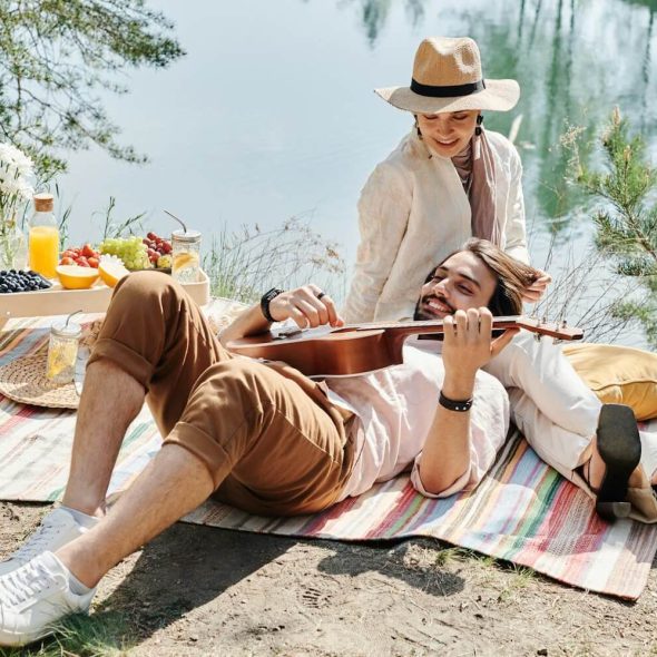 Man resting his head on a woman's lap while playing a guitar, with both sitting beside a lake during a date.