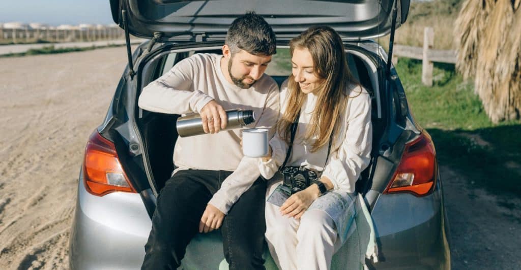 Couple sitting in the trunk of a car, with the man pouring water into a cup held by the woman.