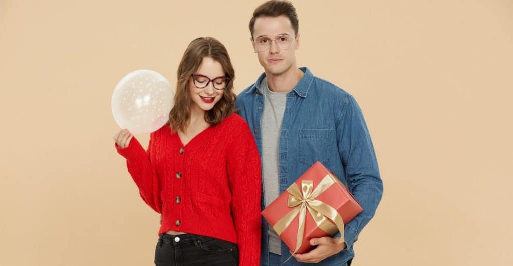 Smiling couple standing together, with the woman holding a white balloon and the man holding a red gift box with a gold ribbon.