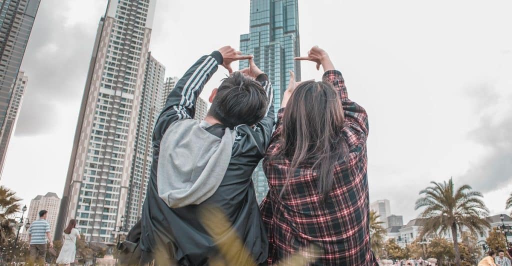 Young couple sitting on grass, making hand camera gestures while facing high-rise buildings.