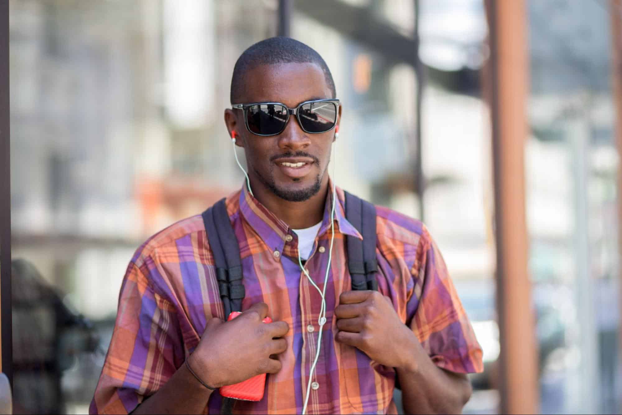Young man in Madras shirt standing on street listening to music from phone. Wearing sunglasses and backpack.
