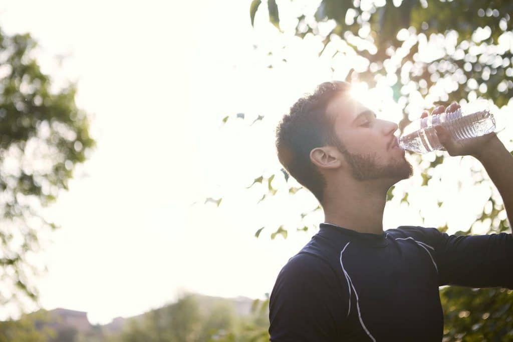 A bearded man in a black shirt drinking water from a plastic bottle with the sun shining behind him.