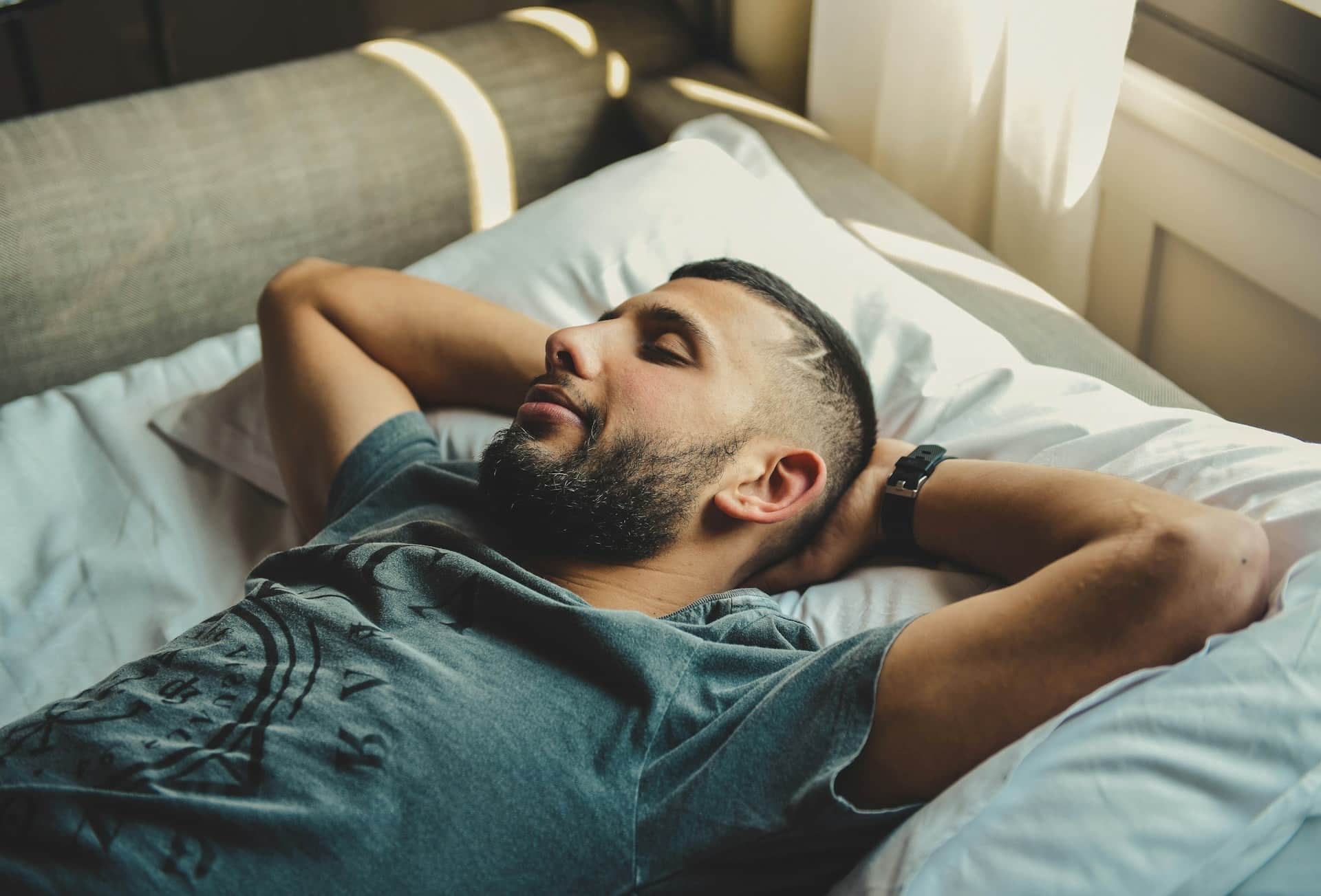 A man resting on a bed with both of his hands beneath his head.