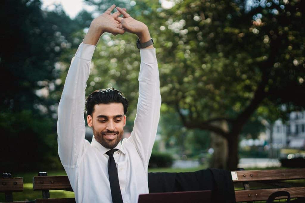 Man in White Dress Shirt and Black Necktie Stretching His Arms Sitting on a wooden bench in a park with a laptop on his lap.
