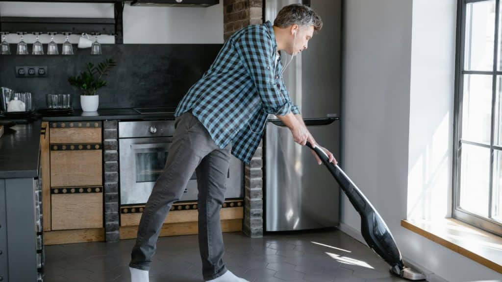 Man in a checkered shirt vacuuming the kitchen floor while wearing wired headphones.