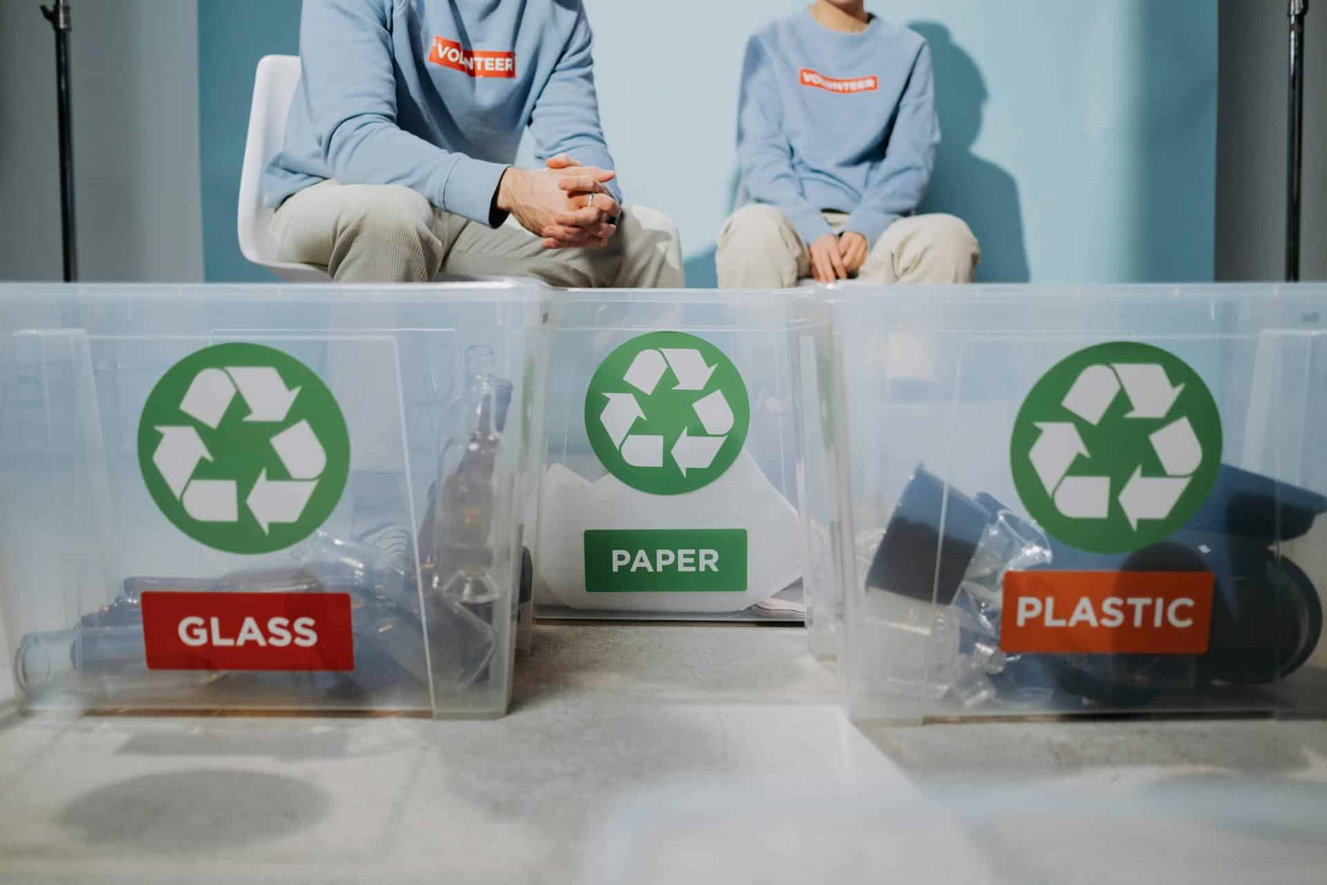 Two people seated on chairs with three plastic recycling bins placed in front of them.