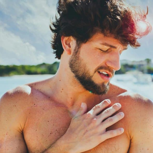 Shirtless man touching his chest while standing on a beach, with water in the background.
