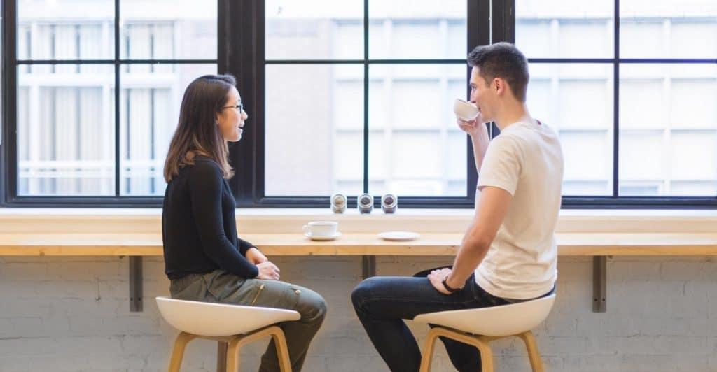 Man and woman sitting on chairs having tea beside a large three-panel window.