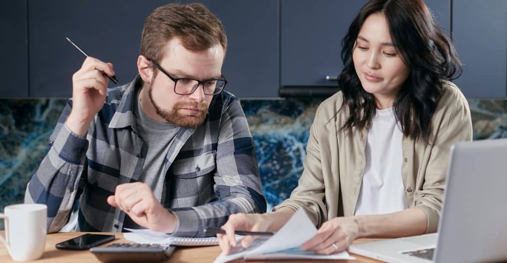 Couple sitting at a desk looking at a paper, with a calculator, phone, mug, and laptop placed in front of them.