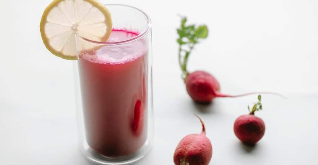 A glass of beetroot juice garnished with a lemon slice, sits next to three small fresh radishes on a white surface.