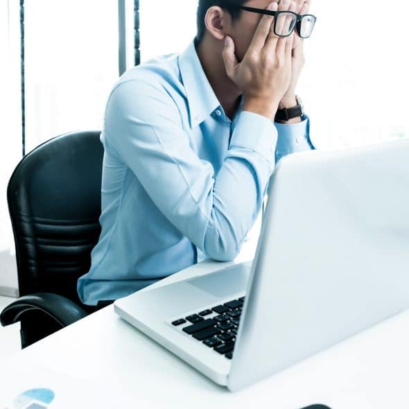 A person wearing a blue shirt and glasses sitting at a desk with a laptop, covering their face with their hands.