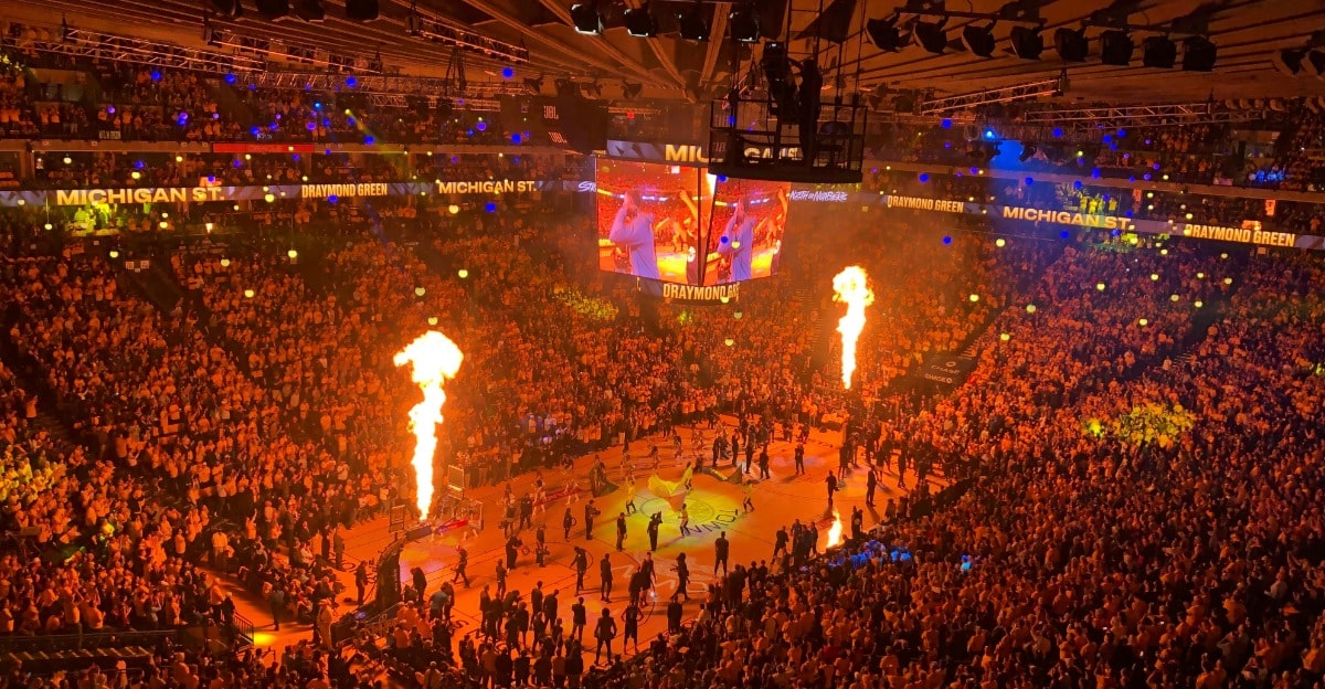 A wide shot of a large indoor arena filled with a crowd of people, with a stage area featuring screens and flames shooting up.