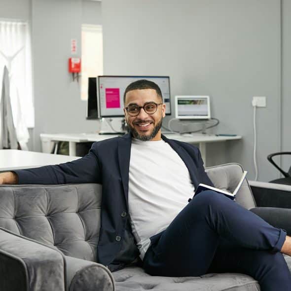 A man with glasses and a beard, wearing a navy blazer, white t-shirt, and navy pants, sitting on a gray couch in an office setting, holding a notebook and pen.