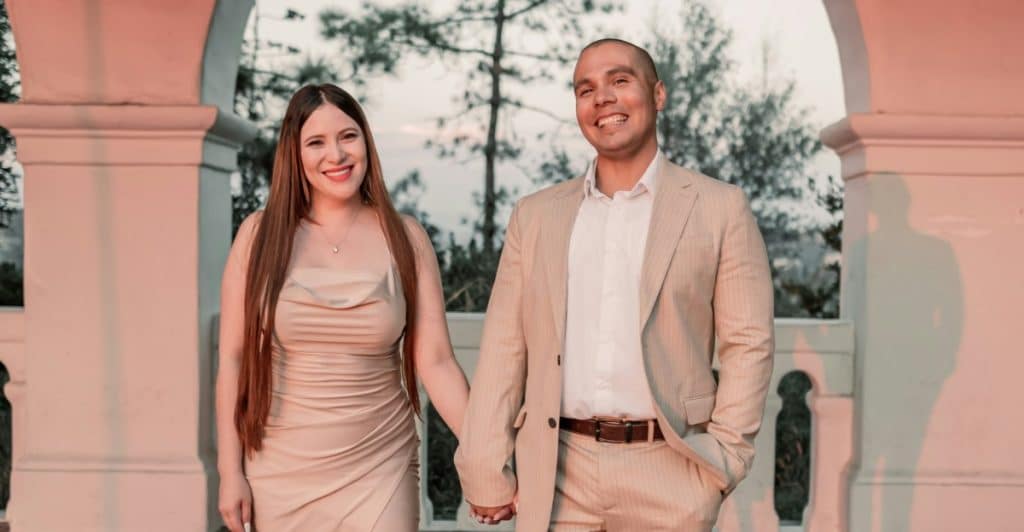 A couple dressed in complementary neutral-toned outfits smiles and holds hands in front of an architectural backdrop.