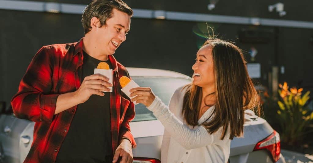 A man and a woman standing outside, sharing food from small paper wrappers.