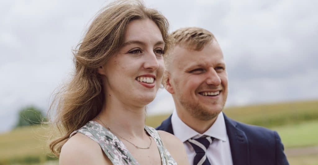 A happy couple poses for a photo outside, dressed in stylish wedding guest outfits.