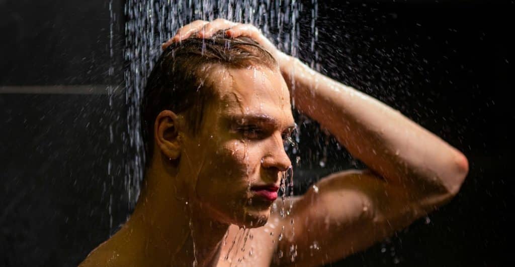 A man standing under a shower, with water hitting his head and shoulders, with closed eyes and water dripping from his face.