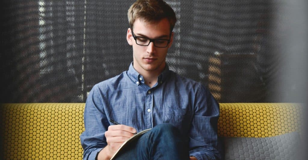 A man wearing glasses and a blue shirt sitting on a yellow couch, writing in a notebook.