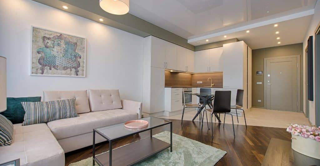 A contemporary living room with a couch, table, rug, and a glimpse of a kitchen area.