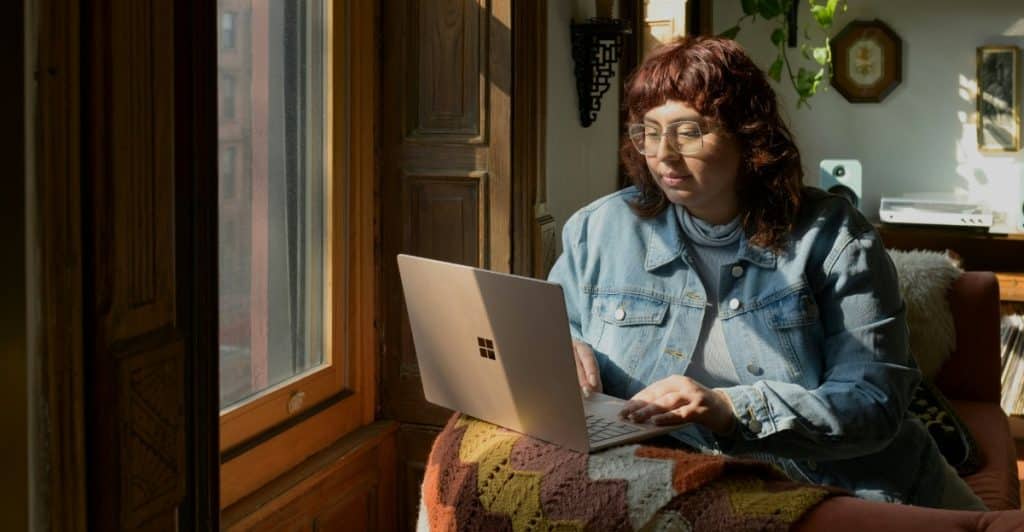 A person with reddish-brown hair and glasses using a laptop in a sunlit room.