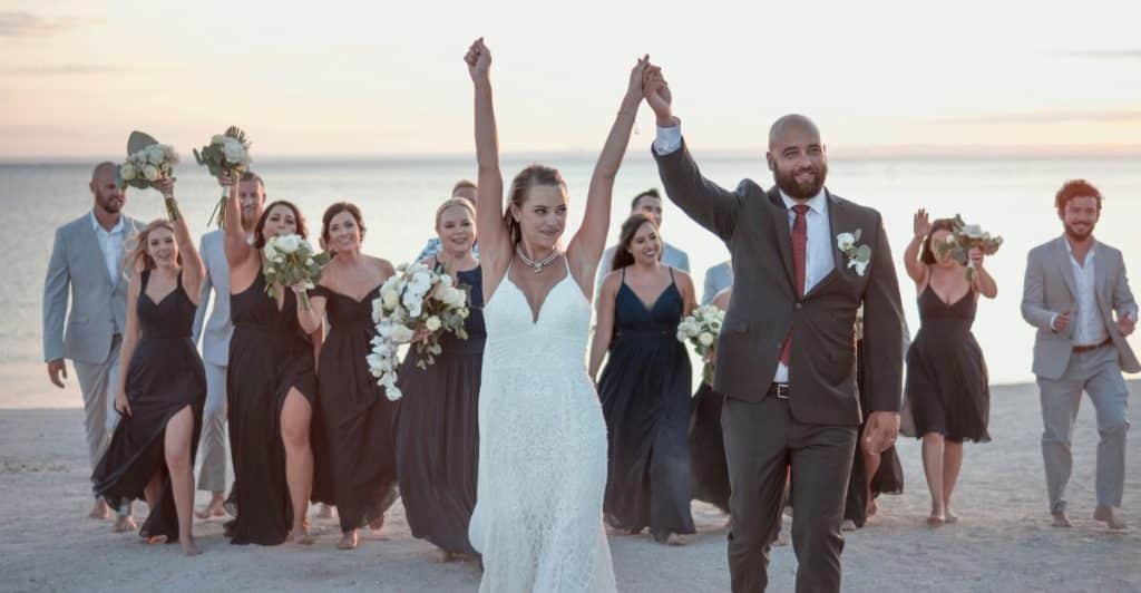 A bride, groom, and their wedding party stand on a beach, celebrating as the sun sets.