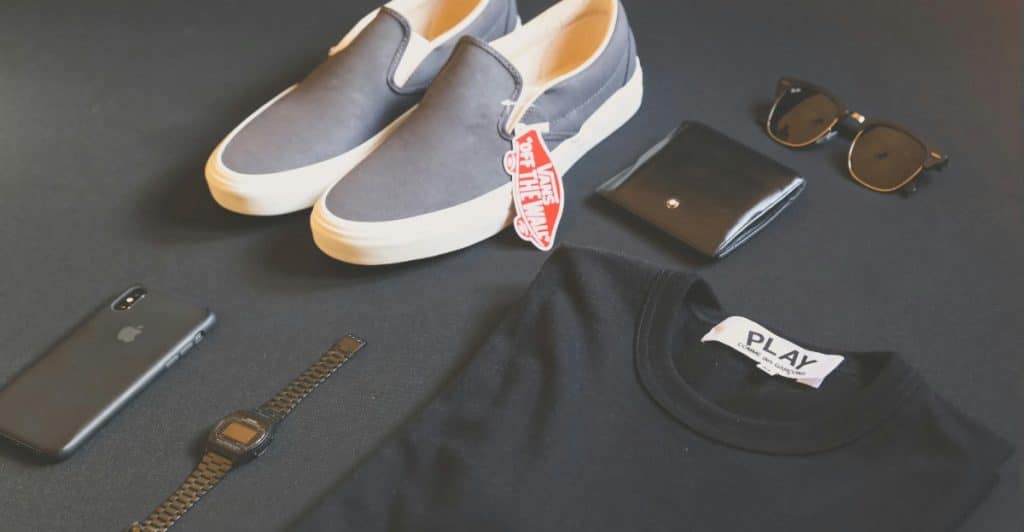 Shoes, a t-shirt, a phone, a wallet, sunglasses, and a watch arranged on a dark surface.