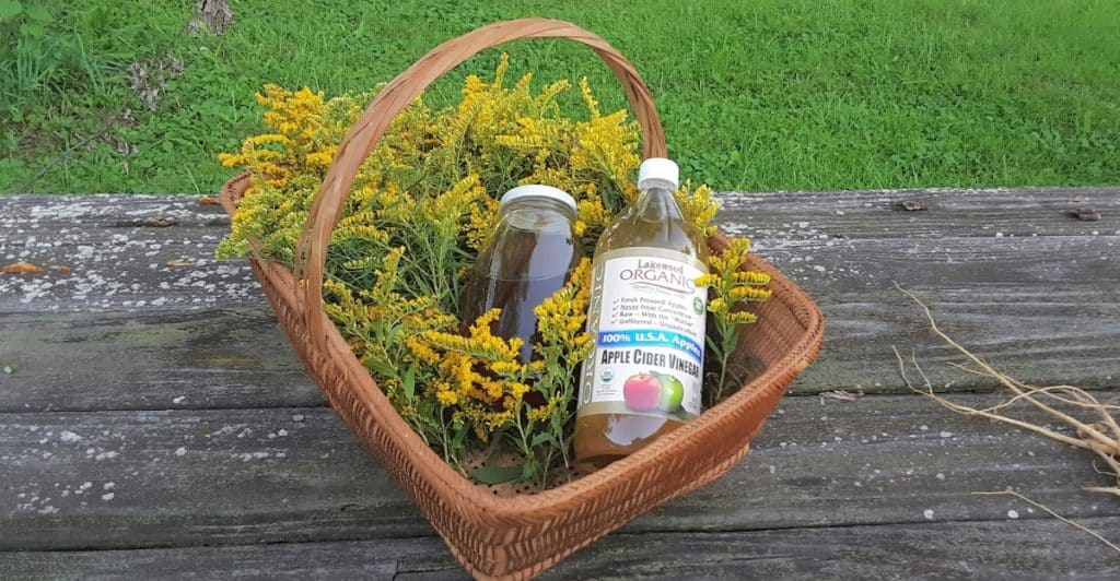 A basket filled with yellow flowers and two bottles, one labeled "APPLE CIDER VINEGAR," are on a wooden surface outdoors.