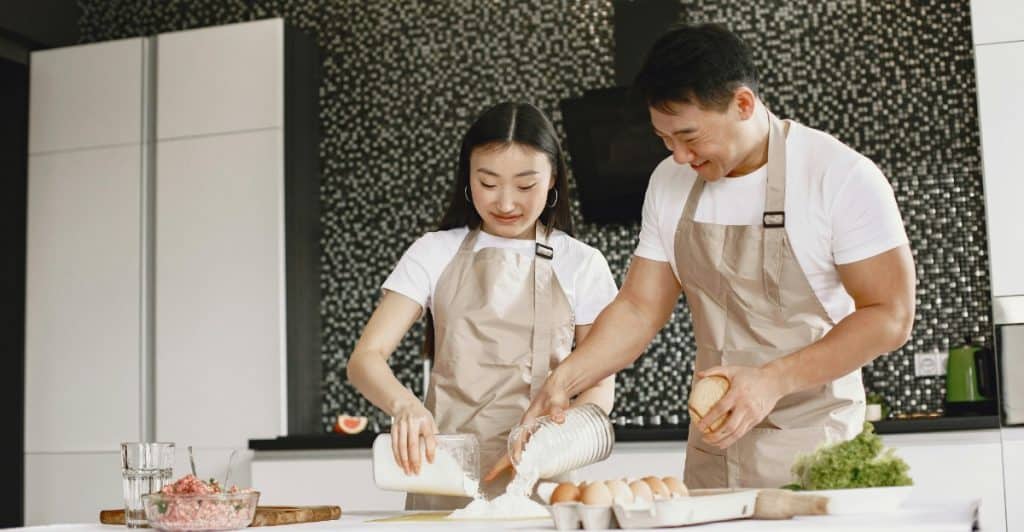 Asian couple in matching shirts and aprons baking together in a kitchen..