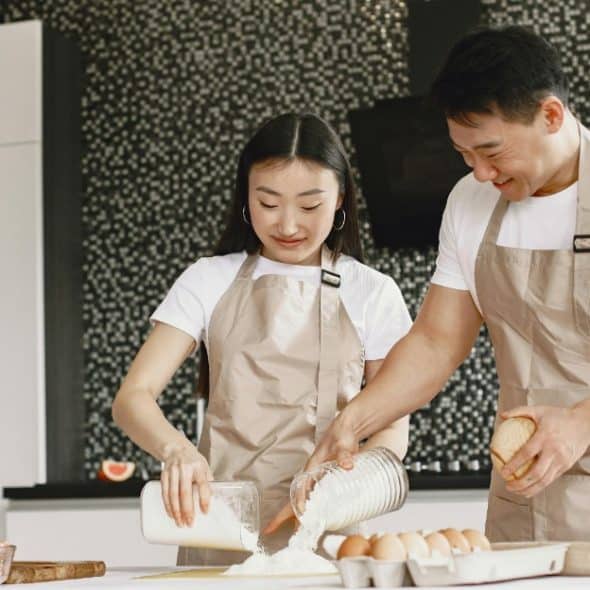 Asian couple in matching shirts and aprons baking together in a kitchen..