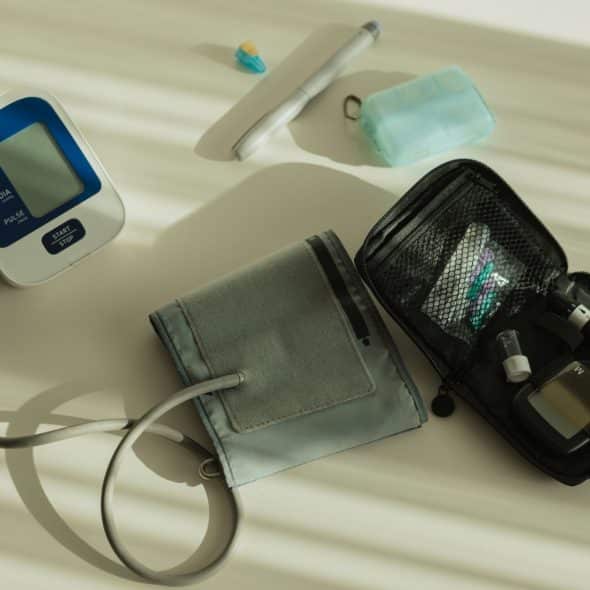 A high-angle shot of a digital blood pressure monitor, a grey blood pressure cuff, a black medical kit, a white pen-like device, and a small blue object.