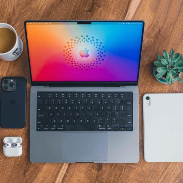 A laptop, smartphone, tablet, earbuds, coffee cup, and small plant are arranged on a wooden table.
