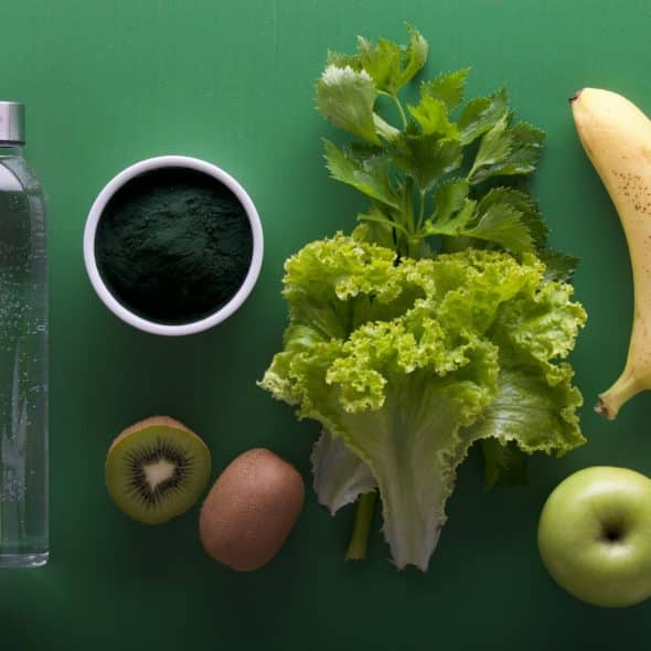 Overhead shot of a metal water bottle, a white bowl of dark green powder, leafy green vegetables, a banana, kiwi fruits, and a green apple arranged on a solid green background.